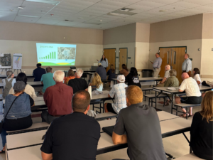 A presenter shows a slide to a seated audience during a community presentation.