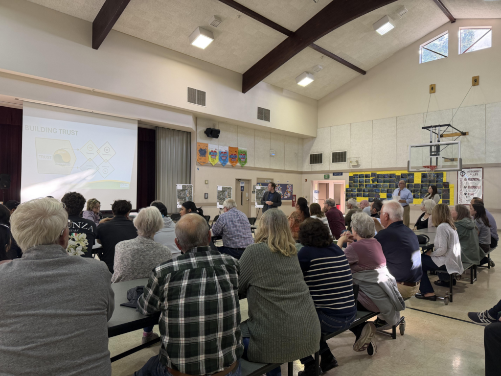 A group of people sit in a community hall, attentively listening to a speaker presenting on "Building Trust" projected on a screen.