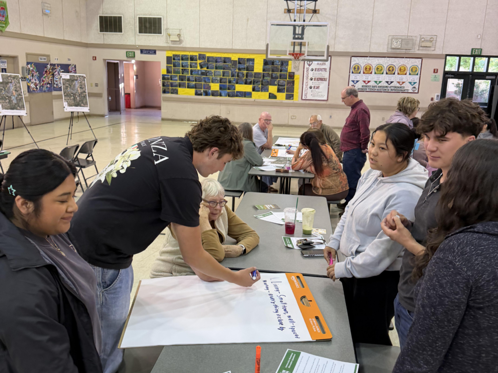 A group of people is gathered around a table in a community hall, engaged in a collaborative activity. A young man writes on a large paper while others watch attentively.
