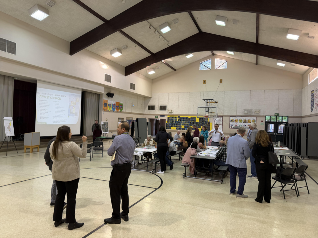 Community meeting in a gymnasium, people engaged in discussions around tables. A large presentation screen displays information.