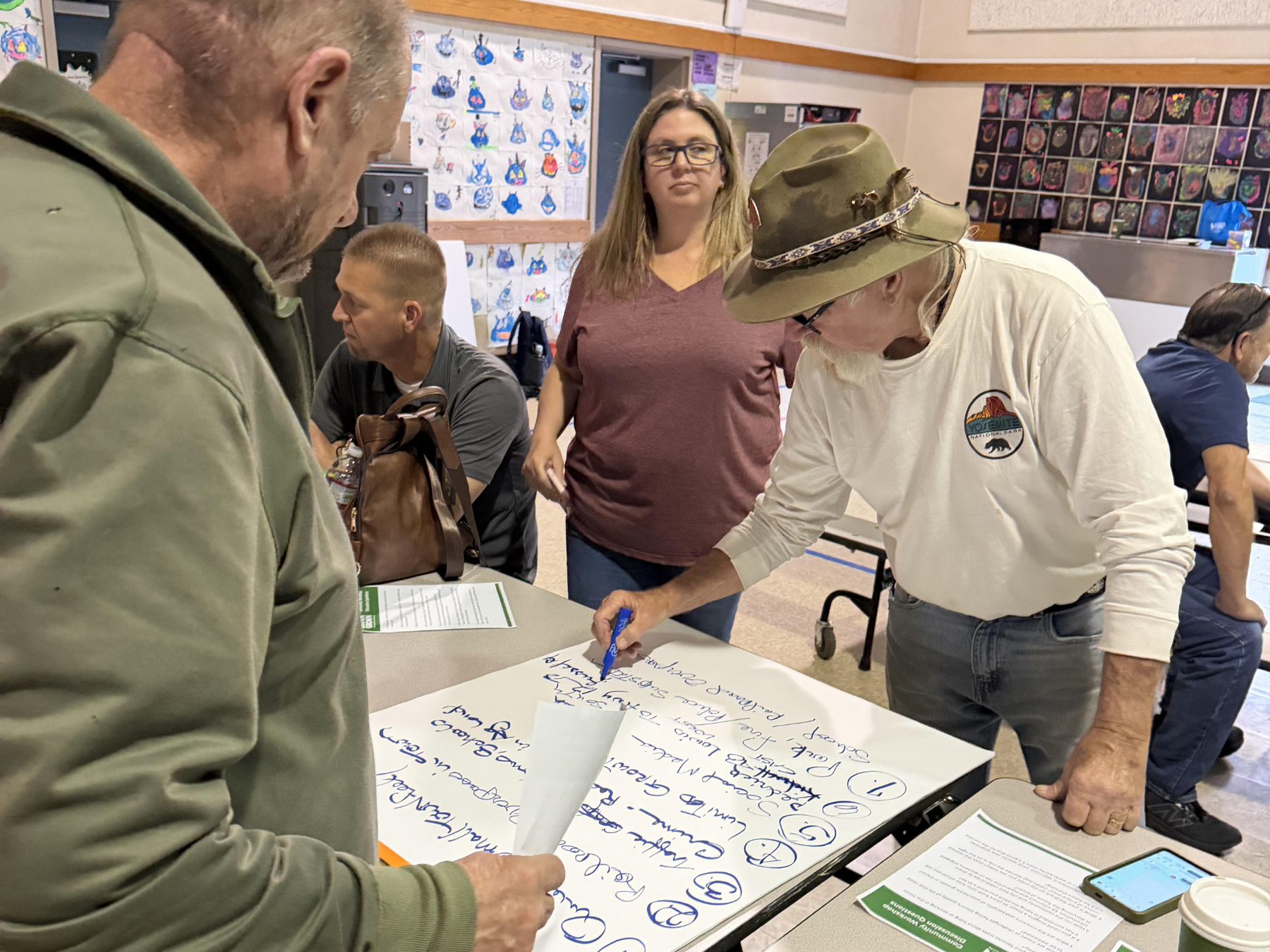 A group of people in a room collaborate around a table covered with notes. One man writes on a paper.