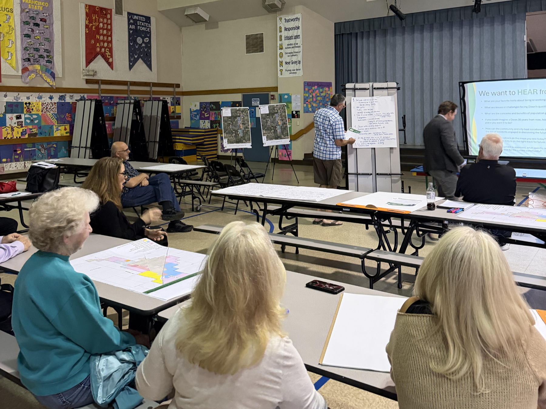 A community meeting in a school gym with several seated attendees facing a presentation. Two men stand near a large screen and flip chart.