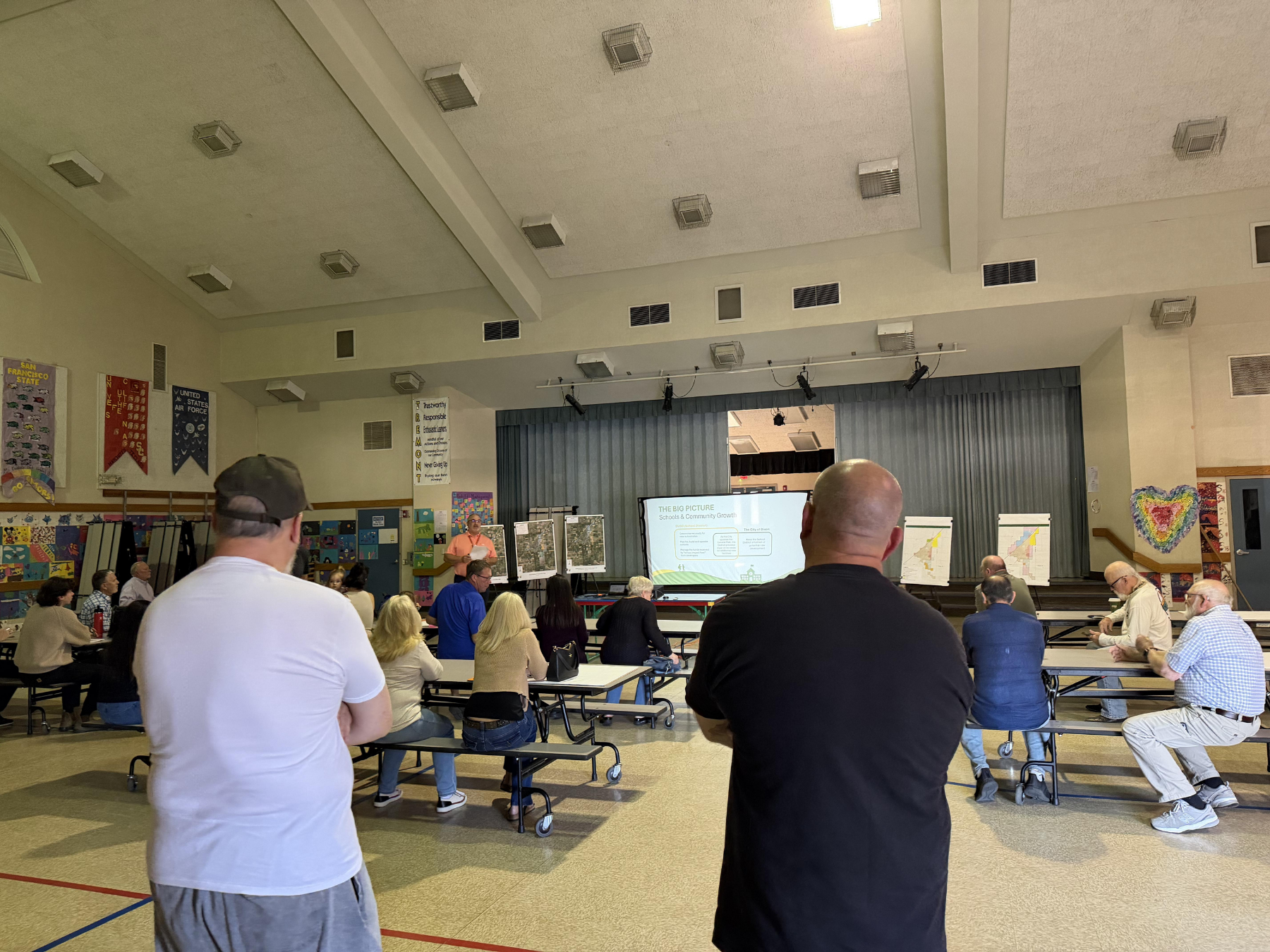 A group of adults sits at cafeteria tables in a school gymnasium, facing a presentation screen displaying information.