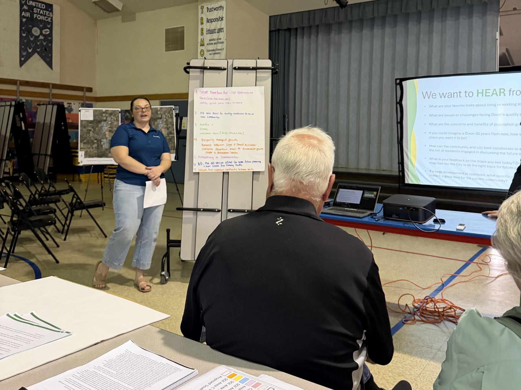 A woman presents at a community meeting in a gym. She stands by an easel with notes, facing attendees seated at tables.