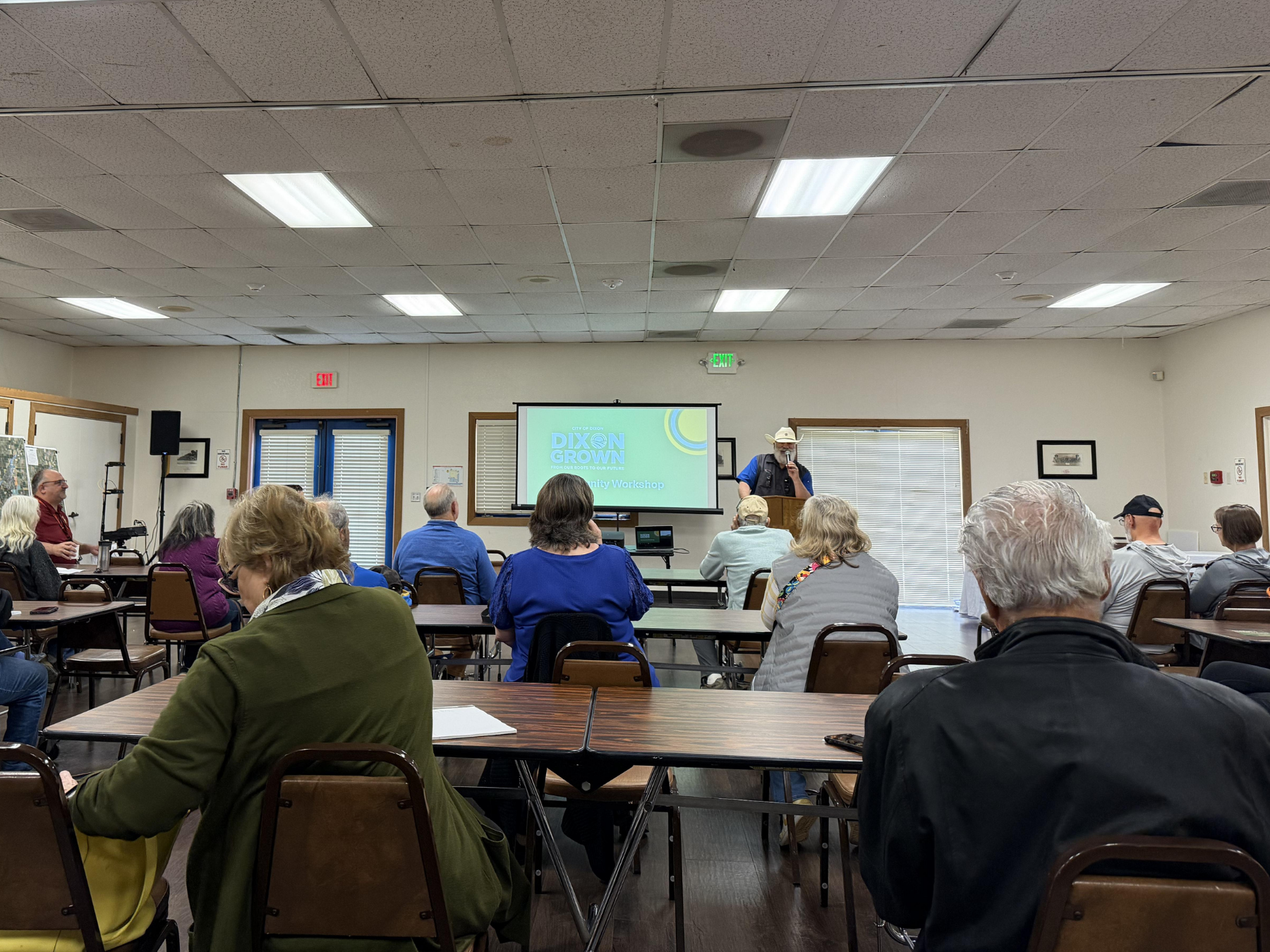 Community meeting with attendees seated at tables, listening to a speaker at a podium.