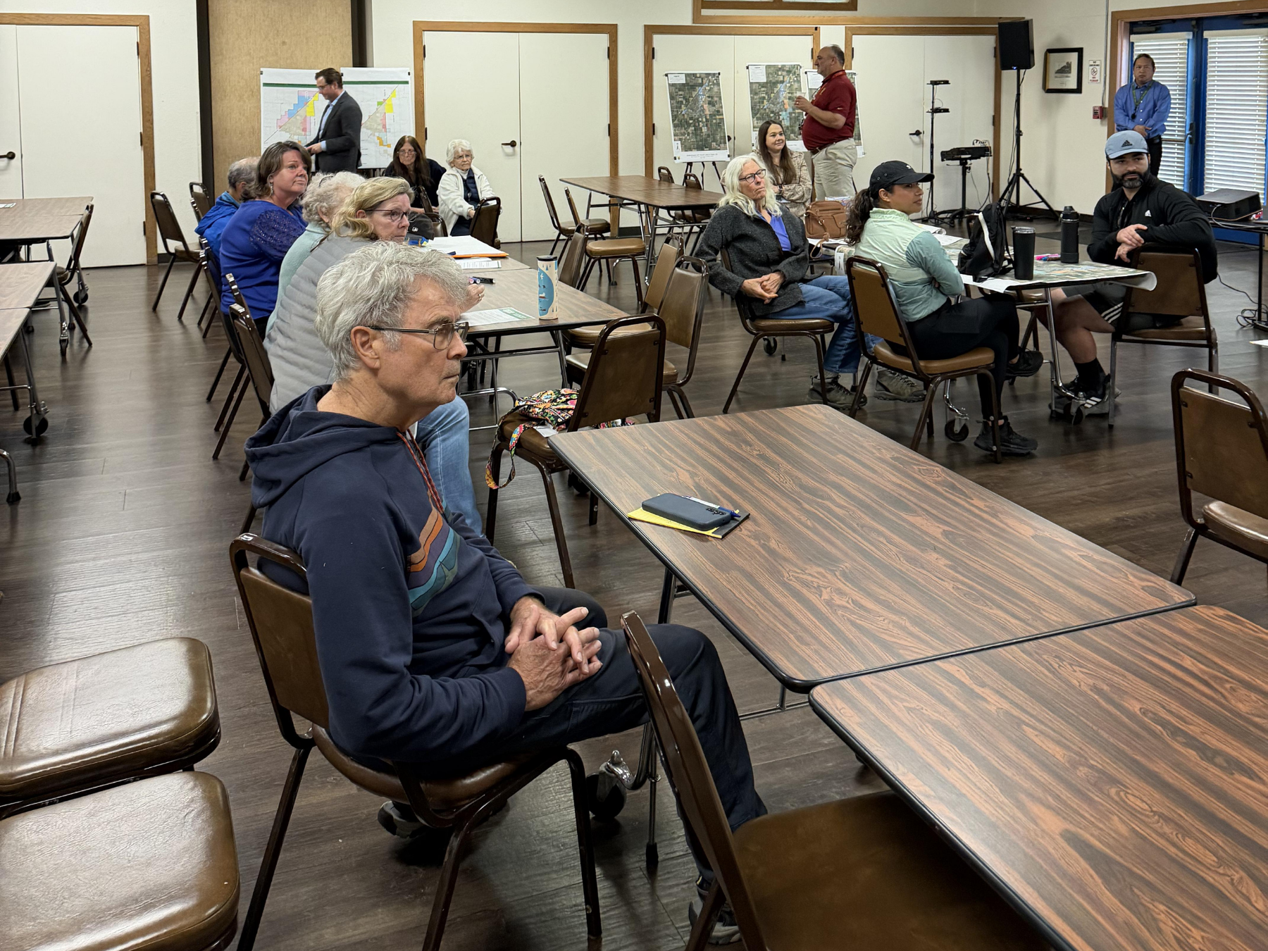 A group of people sits attentively in a room around folding tables, focused on a speaker.