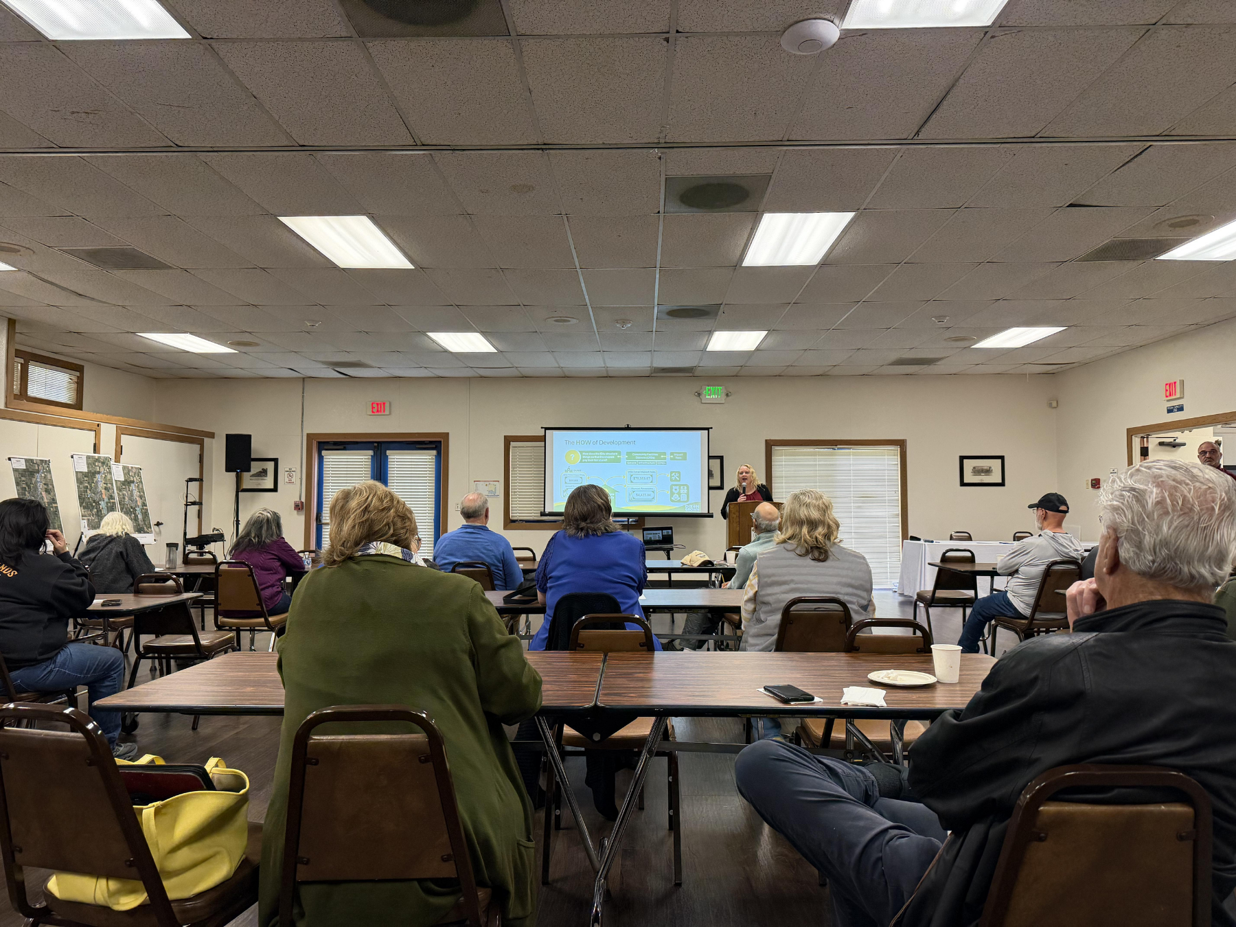 A community meeting in a room with tables and chairs. Attendees face a presenter standing beside a projector screen displaying a slide.