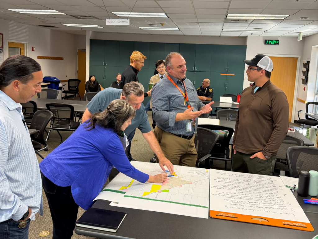 A group of attendees and officials gather around a table to examine and point at a map during the community workshop.