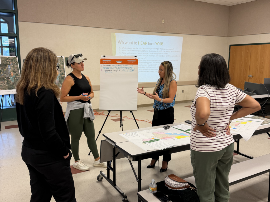 A small group of attendees stand together discussing information on a flip chart and maps at the community workshop.