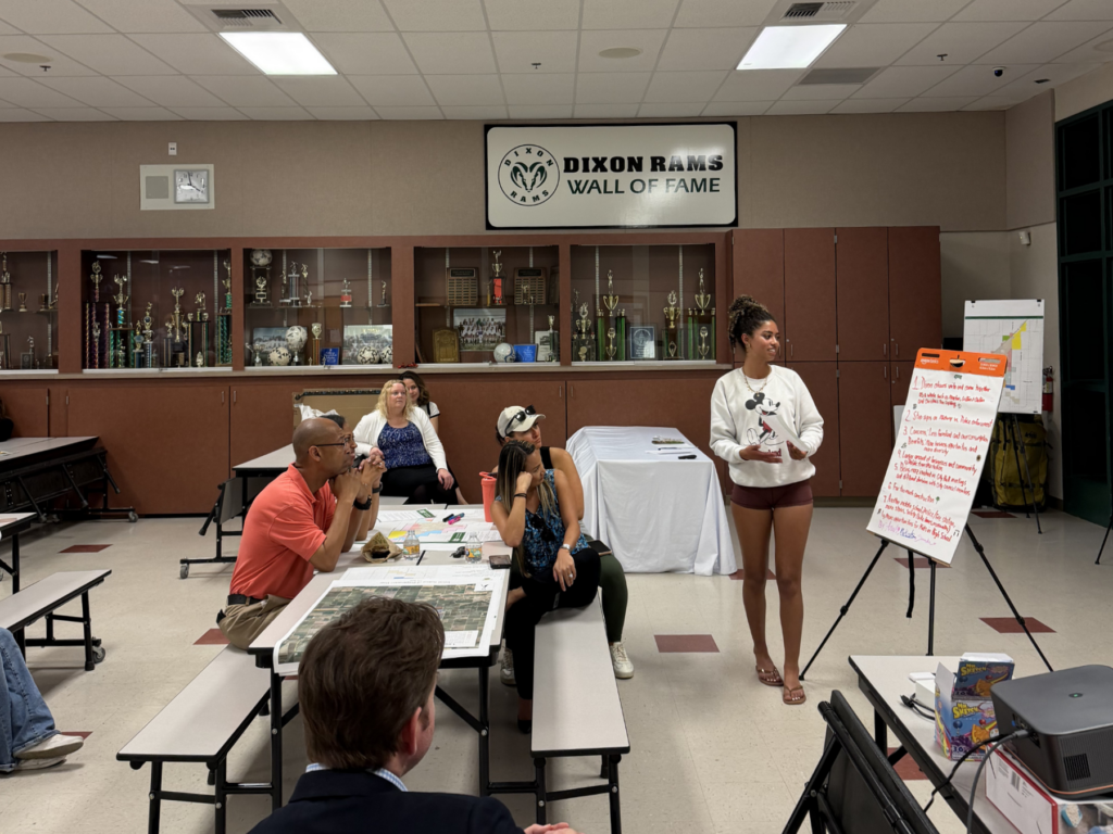 A presenter stands by a flip chart speaking to attendees seated at a table during the community workshop.