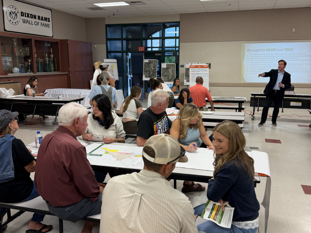 Attendees sit at tables reviewing maps while a presenter speaks in the background at the community workshop.