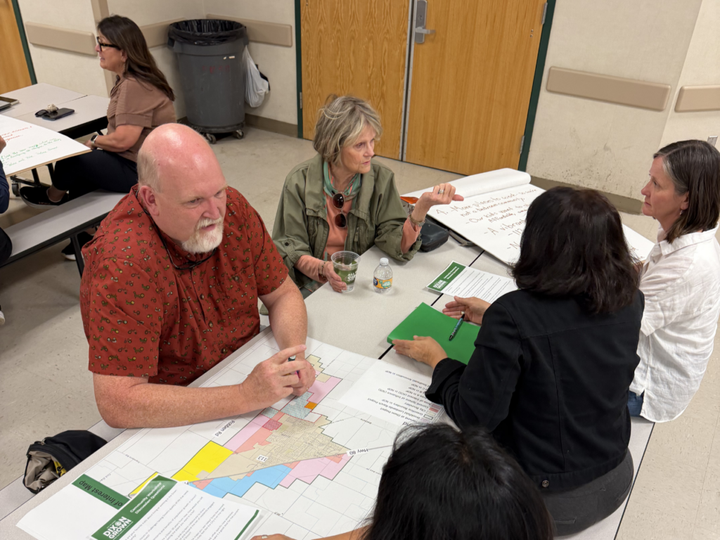 A small group of people examine a colorful map and converse at a table during the community workshop.