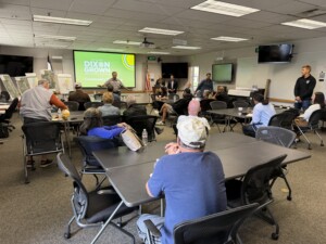 Community members seated at tables in a room facing slide deck with "Dixon Grown" text and presenter.